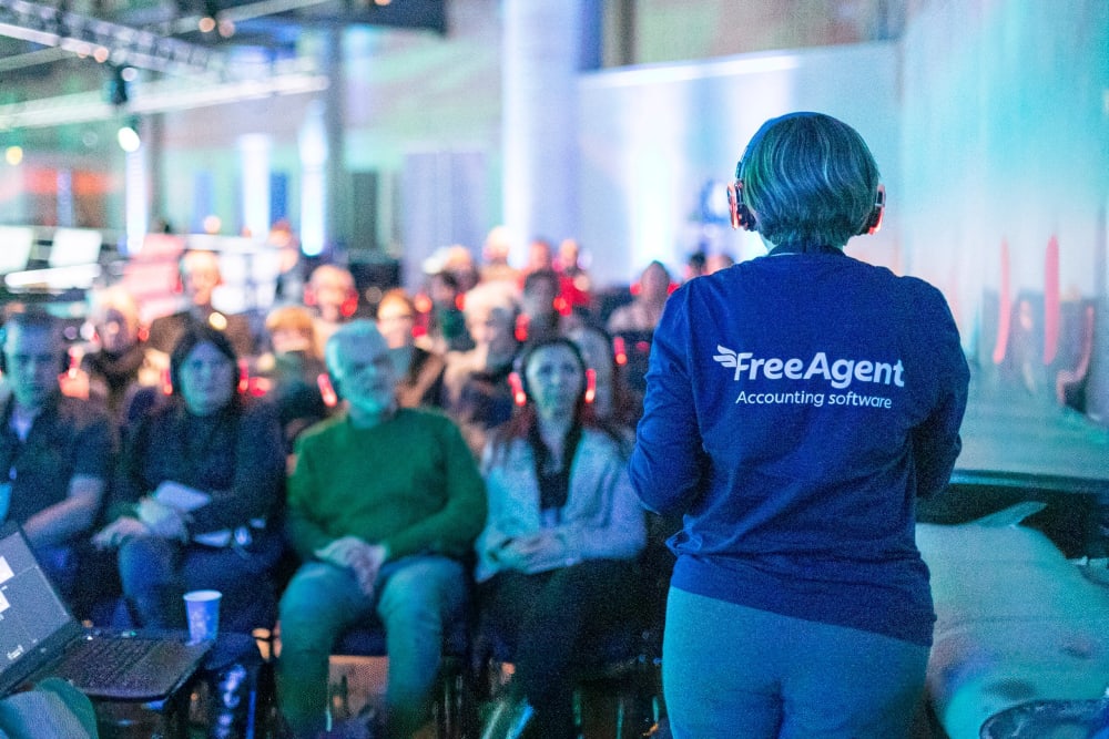 A person wearing a FreeAgent t-shirt stands with their back to the camera. They are presenting to a group of event attendees in front of them. The audience and the speaker are wearing headphones