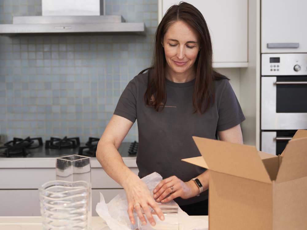 Two women wearing grey T shirts, Kath Woods and Zoë Miller of All About The Move, are looking at a mobile screen. There are cardboard boxes stacked beside them.