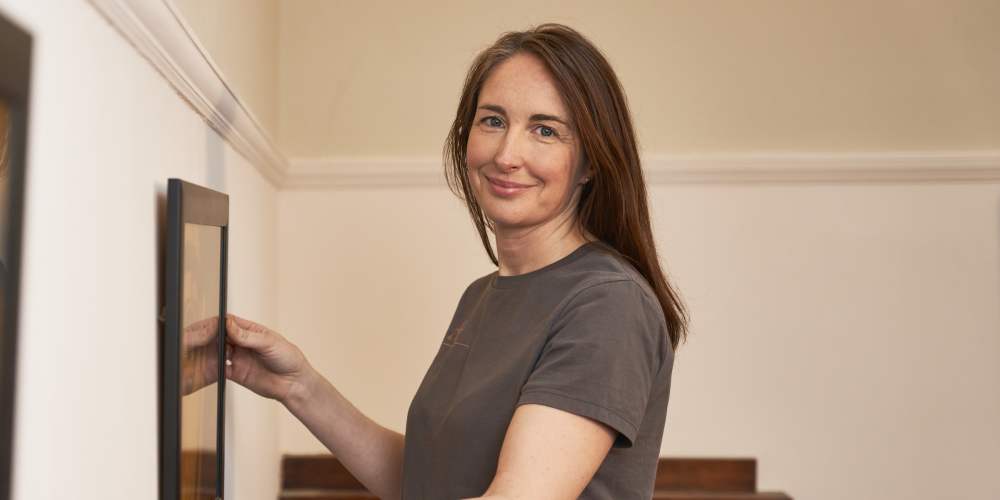 A woman, Zoë Miller, is placing picture frames on a wall during a house-move.