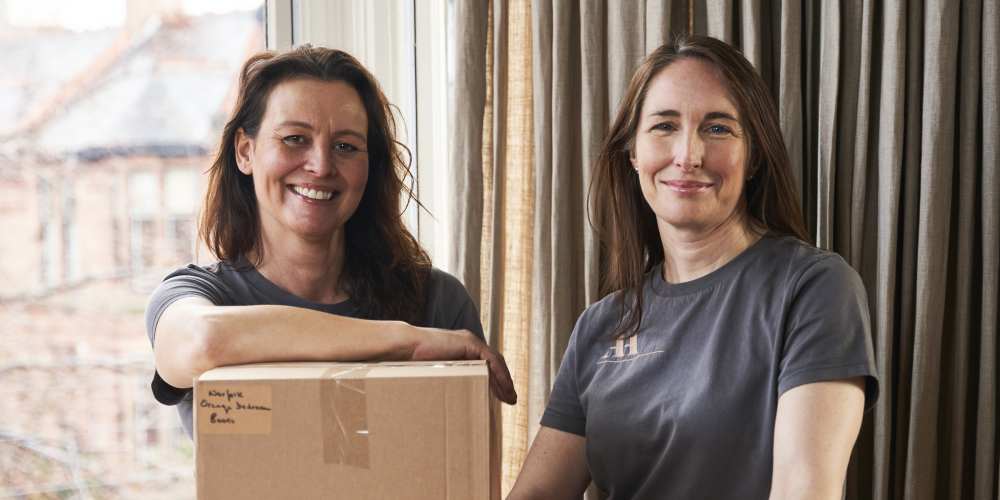 Two women, Kath Miller and Zoë Woods, stand in front of a window with grey curtains. Kath on the left is leaning her elbow on a cardboard box, both are smiling at the camera.