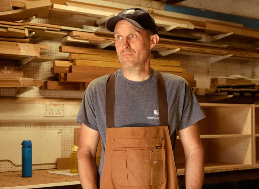 A man wearing an carpenter's apron stands in front shelves with wood and tools on them.