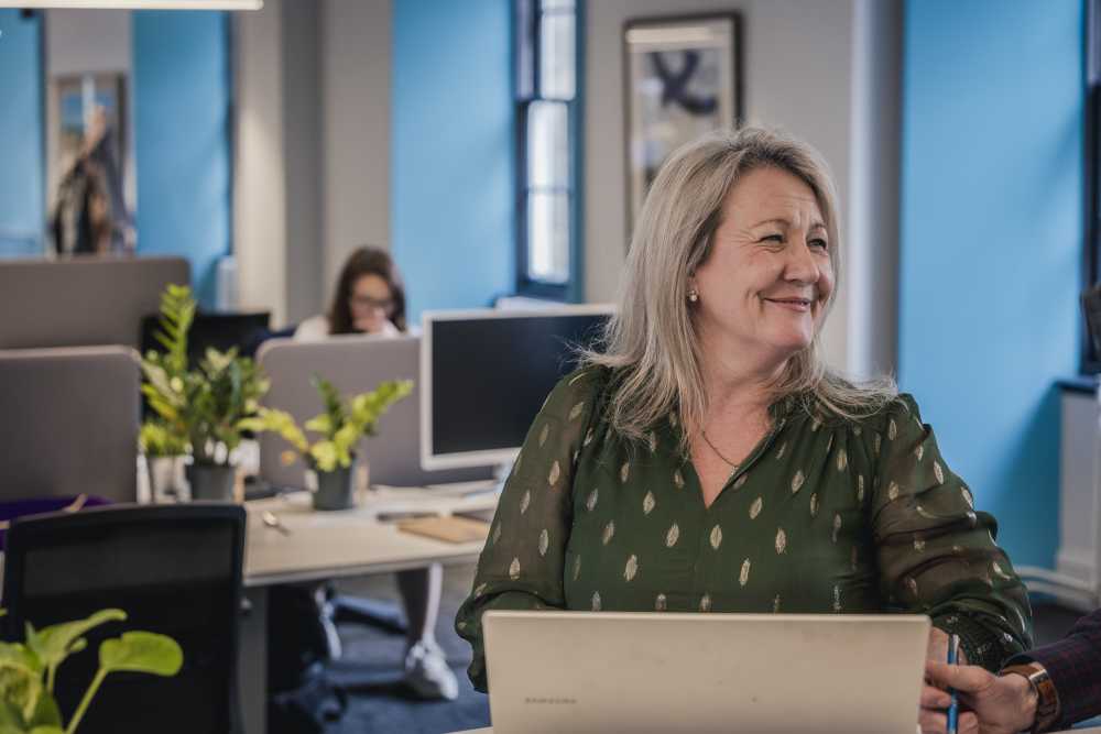 A smiling woman in a green blouse sits at a laptop