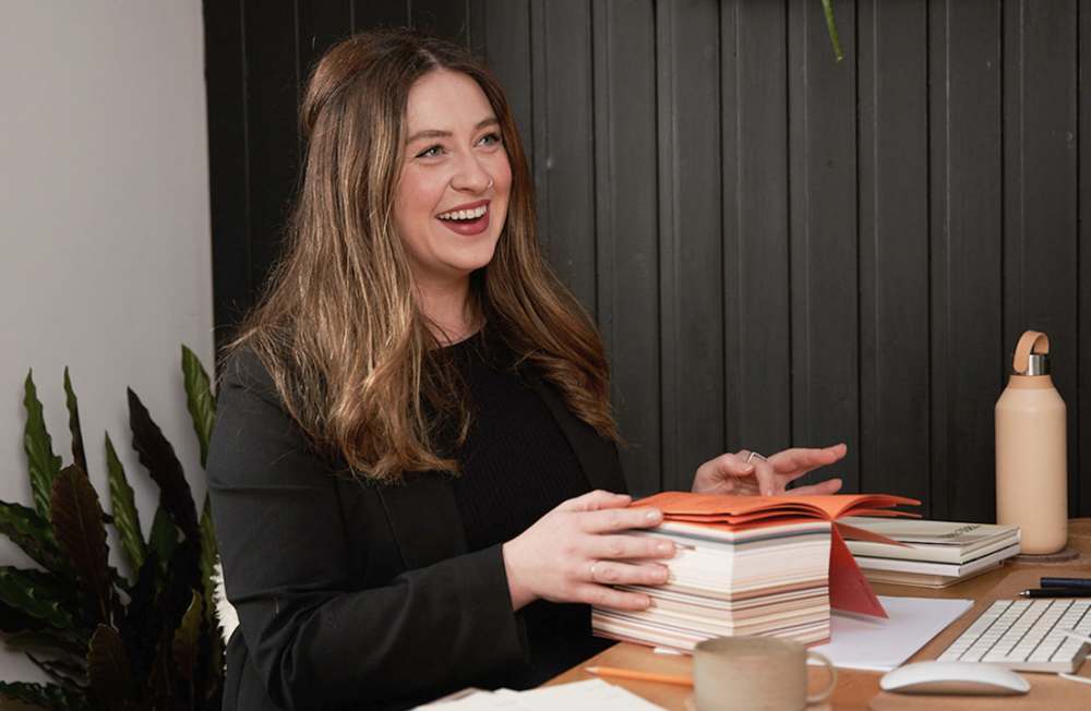 Lisa, smiling at her desk, holds a thick stack of colourful paper samples.