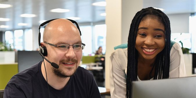 Two members of the FreeAgent support team, one male wearing a telephone headset, the other female and standing to his right, are smiling and having a conversation while looking at a computer screen.